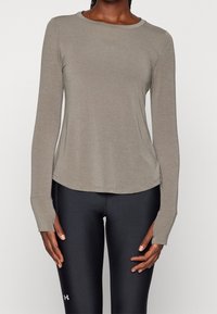 Dlouhý rukáv, světle hnědý sportovní top vyrobený z měkkého materiálu, s kulatým výstřihem a zakřiveným lemem, nošený s černými legínami.