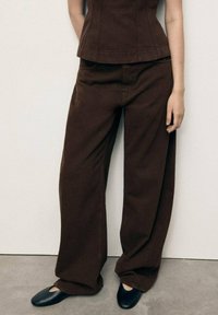 Brown wide-leg trousers made of cotton, featuring a relaxed fit, side pockets, and a smooth texture. Paired with black flat shoes.