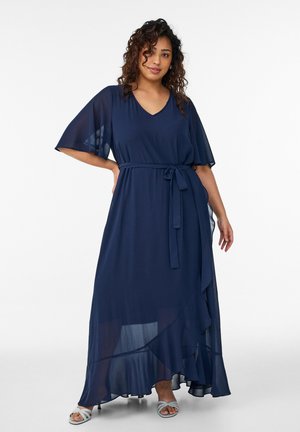 Vrouw draagt een marineblauwe maxi-jurk met V-hals, doorschijnende fladdermouwen en een tailleceintuur, gecombineerd met lichtblauwe sandalen met bandjes.