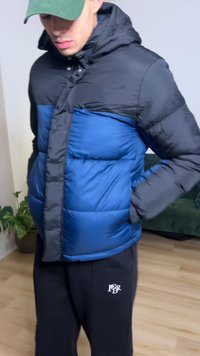 Piumino con davanti blu e maniche nere, dotato di cappuccio, chiusura con zip e polsini elastici. Indossato sopra pantaloni della tuta neri.