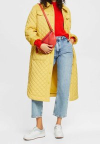 Gesteppter gelber Mantel, roter Strickpullover, hellblaue gekürzte Jeans, weiße Turnschuhe mit grauen Akzenten und kleine rote Umhängetasche.