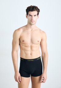 Boxeri scurți negri din bumbac, cu un logo galben Calvin Klein pe banda de talie, design ajustat și textură netedă. Modelul stă pe un fundal neutru.