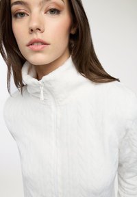 Biała polarowa kurtka z zamkiem błyskawicznym i wysokim kołnierzem, charakteryzująca się pikowanym materiałem, gładkim zamkiem oraz dopasowanym krojem. Brak widocznych wzorów lub akcentów.