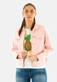 Chaqueta de denim rosa claro con botones dorados, dos bolsillos en el pecho, llevada sobre una camiseta blanca con un gráfico de piña, combinada con unos jeans azules.