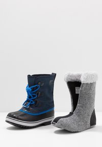 Wodoodporny czarny gumowy śniegowc z niebieskimi akcentami i sznurówkami po lewej; szary filcowy but z białym futerkiem po prawej.