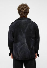 Zwarte, fluffy, oversized fleecejas met een gestructureerd oppervlak, voorzien van een kraag en lange mouwen. De achterzijde toont subtiele patroonvariaties.