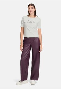 Femme portant un T-shirt gris clair avec des motifs de flacon de parfum, un pantalon en cuir violet foncé ample, et des baskets blanches à plateforme, se tenant droite.