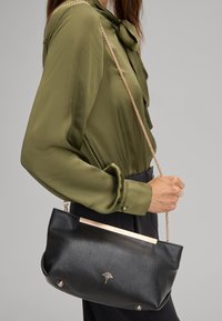 Borsa nera textured con dettagli dorati e tracolla a catena, tenuta da una persona che indossa una blusa verde. Presenta un dettaglio del logo nella parte anteriore.