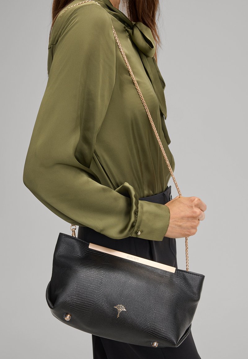 Borsa nera textured con dettagli dorati e tracolla a catena, tenuta da una persona che indossa una blusa verde. Presenta un dettaglio del logo nella parte anteriore.