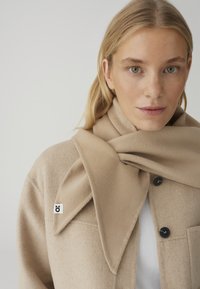 Blonde Frau trägt beigen Wollmantel und passenden Schal, mit grünen Augen und neutralem Gesichtsausdruck vor einfarbigem Hintergrund.