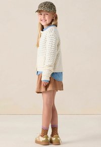 Fille souriante portant une casquette à imprimé léopard, un pull en tricot crème sur une chemise bleue, une jupe en velours côtelé marron, des chaussettes à motif de cœur et des baskets beiges, debout.