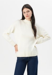 Cremfarbener Strickpullover mit hohem Rippenkragen und gerippten Bündchen; entspannte Passform, weiche Textur und einfarbiges Muster. Getragen mit dunkelblauen Jeans.