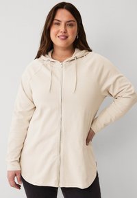 Beige Kapuzenjacke mit Reißverschluss, langen Raglanärmeln und abgerundetem Saum. Hergestellt aus weichem Stoff mit Kordelzug-Details am Hals.
