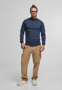 Marinblå zip-up tröja med långa ärmar och ribbade muddar, i kombination med beige cargobyxor med flera fickor och beiga sneakers.