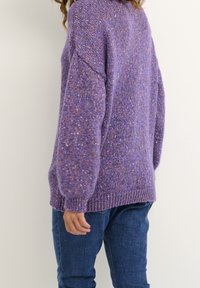 Lila stickad tröja med ett texturerat mönster, lös passform och ribbade manschetter. Matchad med blå jeans, sedd bakifrån.