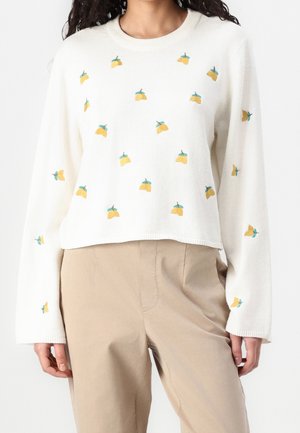 Hvid sweater med lange ærmer med lille gul frugtbroderi kombineret med beige bukser med høj talje, vist fra nakke til hofter.