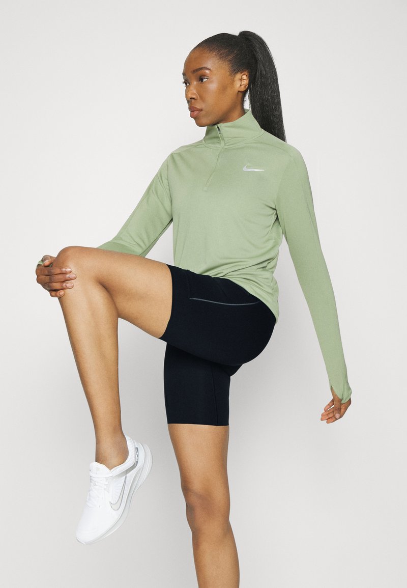Camiseta Nike de manga larga en verde claro, con media cremallera y logo blanco, combinada con pantalones cortos de ciclismo negros y zapatillas deportivas blancas.