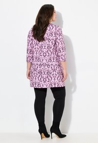 Tunic rosa a fantasia con maniche a tre quarti, abbinata a leggings neri e stivaletti neri con tacco alto. Sfondo neutro.