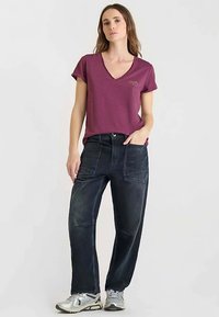 V-hals, bordeauxrode T-shirt met korte mouwen, gecombineerd met donkerblauwe, losse spijkerbroek met zakken, gedragen met lichtgrijze sportschoenen.