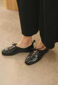 Scarpe nere stringate in pelle lucida con design a punta quadrata e linguetta sul tallone, indossate con pantaloni neri larghi su una superficie chiara.