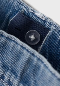 Jeansmidja med en genomskinlig plastknapp, mörkblåt tyg under, med synliga sömmar och fransdetaljer längs kanten.