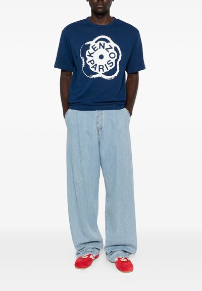 Mand iført navyblå Kenzo Paris t-shirt, lyseblå vide jeans og røde ruskindssko, står med hænderne i lommerne.