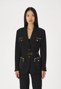 Blazer noir avec boutons dorés, poches zippées et une ceinture avec logo. Coupe ajustée avec manches longues et textures de tissu contrastantes.