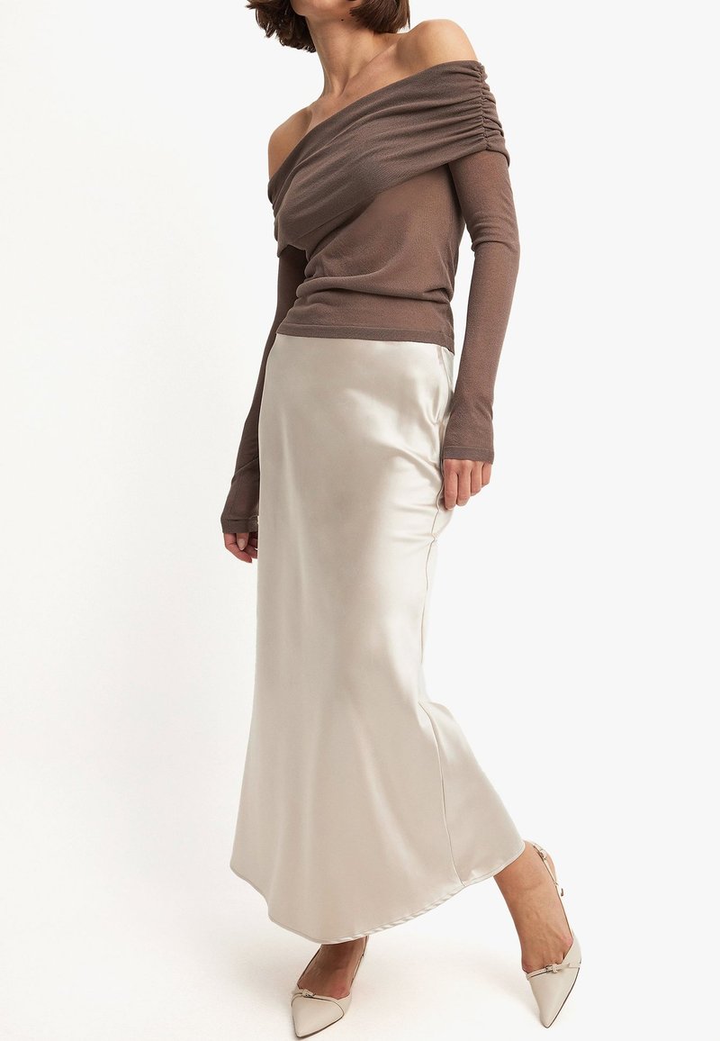Een bruine off-shoulder top met lange mouwen, gecombineerd met een vloeiende satijnen crèmekleurige rok. De schoenen zijn pointed met een subtiele accentdetail.