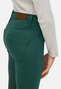 Jeans de mezclilla verdes de talle alto, con un bolsillo trasero y una etiqueta de marca de cuero. Textura suave y diseño ajustado.