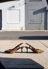 Par leopardprintede højhælede slingback-sko placeret på en fortov med en gade og bygning i baggrunden.