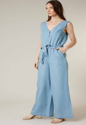 Femme debout portant une combi-pantalon sans manches bleu clair à jambes larges avec ceinture nouée à la taille et sandales à talons beiges, regardant sur le côté.