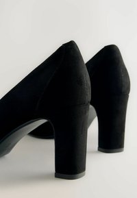 Zapatos de tacón alto de ante negro con un tacón voluminoso, textura suave y un diseño elegante. Incluye detalles de costura sutiles.