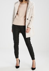 Cappotto grigio chiaro con texture e colletto alto, top beige con scollatura dettagliata, pantaloni skinny neri e scarpe con tacco alto nere.