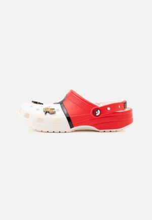 Hvide og røde slip-on klipklappere med ventilationshuller og dekorative vedhæng på tåområdet, med sort rem og tekstureret sål.