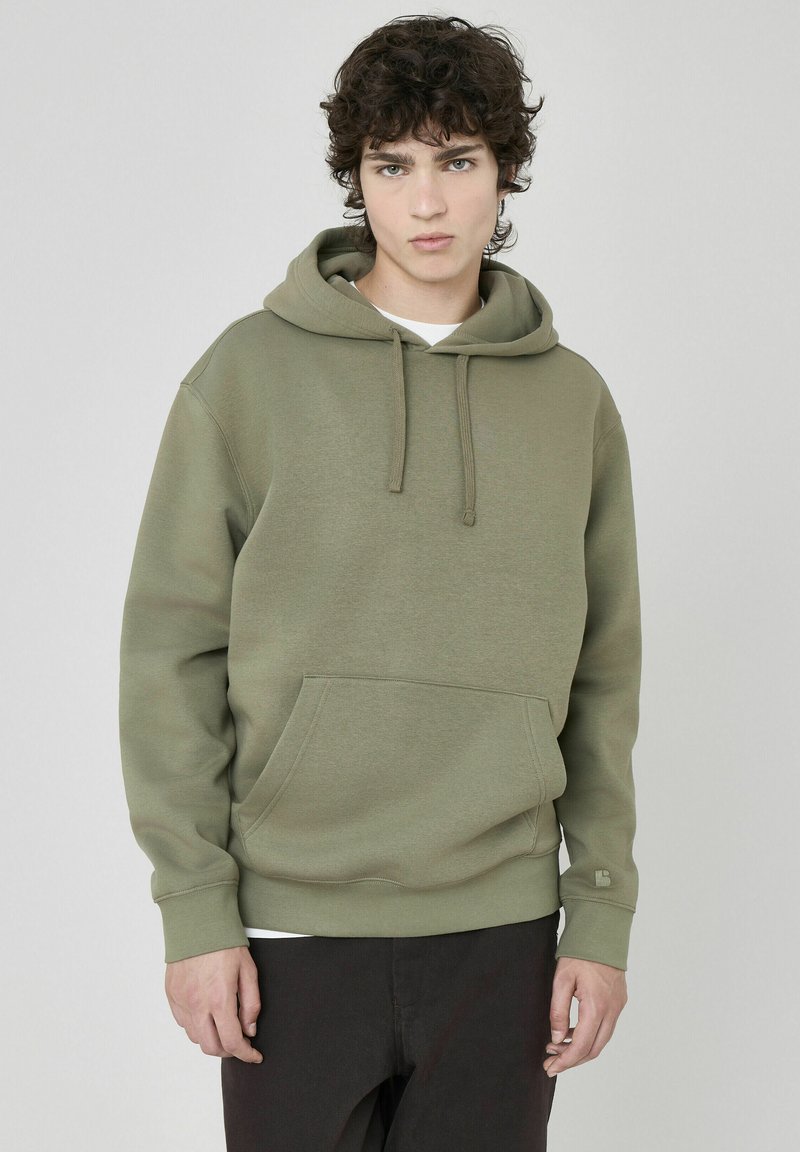 Sweat-shirt à capuche vert olive en tissu doux et texturé, avec une poche kangourou à l'avant et des poignets côtelés. Détail de logo minimaliste sur la manche.