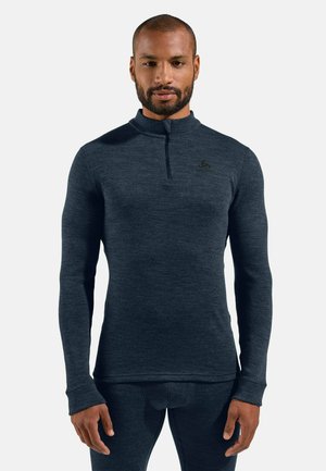 Mann trägt ein eng anliegendes dunkelgraues Langarmshirt mit kurzem Reißverschluss am Hals und passende Hose, steht vor einem einfarbigen Hintergrund.