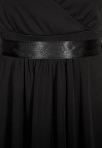 Musta mekko, jossa on tekstuuri ja sileä, kiiltävä musta satiinivyö vyötäröllä. Hame on laskostettu lisädetailin vuoksi.