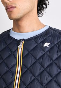Navy quilted jacket with a zip closure featuring a multicolored striped accent and logo detail, complemented by a light blue ribbed collar.