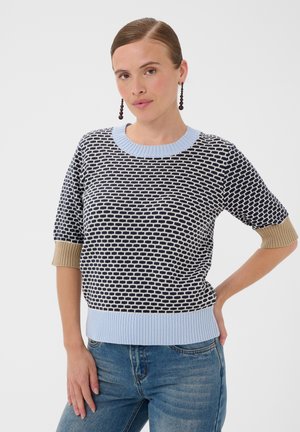 Femme portant un pull à manches courtes noir et blanc à motif avec une finition bleue et des poignets beiges, un jean bleu, et de longues boucles d'oreilles sombres perlées.