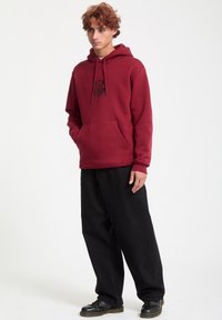 Sudadera roja con bolsillo frontal y diseño gráfico, combinada con pantalones anchos negros y zapatos negros, exhibida sobre un fondo liso.