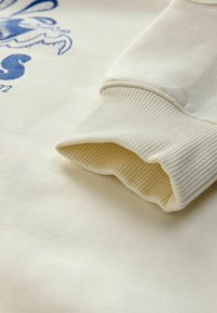 Sweatshirt en coton crème avec des poignets côtelés, une texture lisse et un imprimé graphique bleu à l'avant. Gros plan sur le détail de la manche affiché.