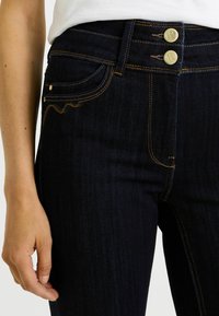 Jeans de mezclilla azul oscuro con cintura alta, dos botones delanteros y detalles de costura sutiles en los bolsillos. La tela tiene una textura suave.