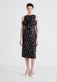 Black sleeveless dress with pink floral pattern, featuring a large bow at the neckline, fitted silhouette, and knee-length hem. Worn with pink clogs.