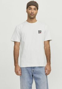 Hvid bomulds T-shirt med korte ærmer, prydet med et lille sort og rødt logo på venstre bryst, sammen med lyseblå jeans.