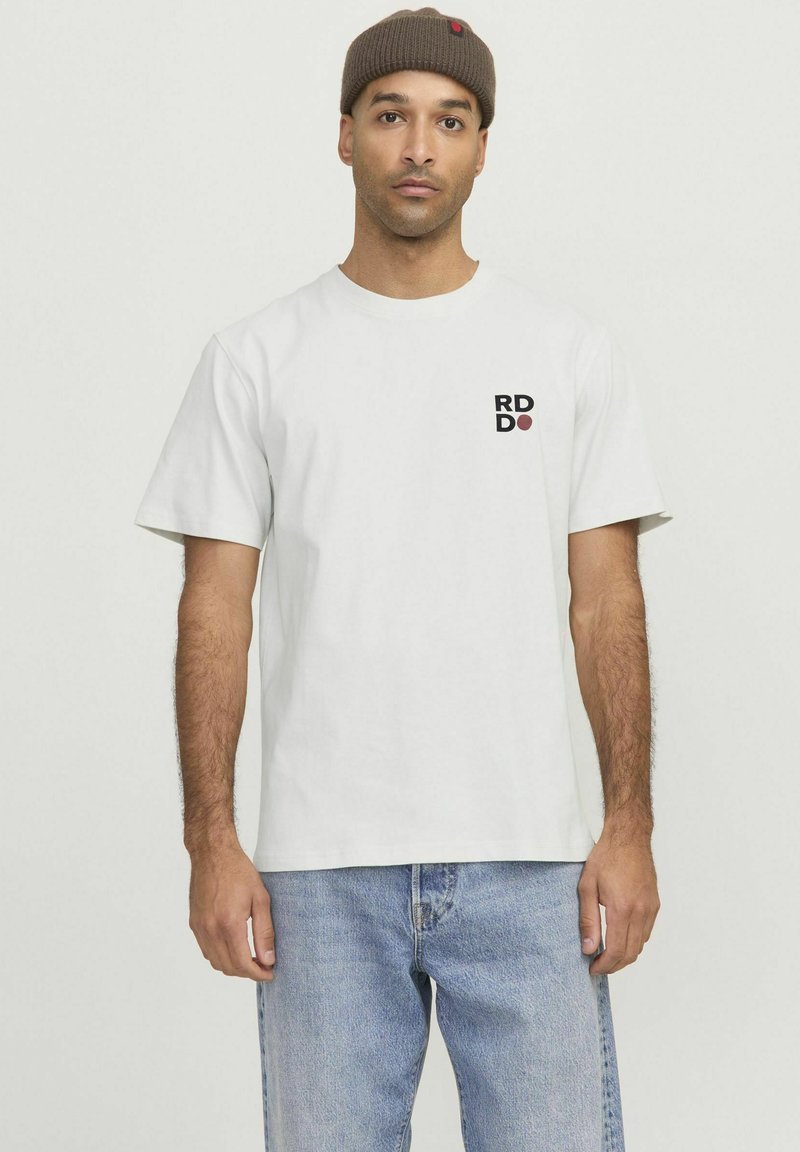 Hvid bomulds T-shirt med korte ærmer, prydet med et lille sort og rødt logo på venstre bryst, sammen med lyseblå jeans.