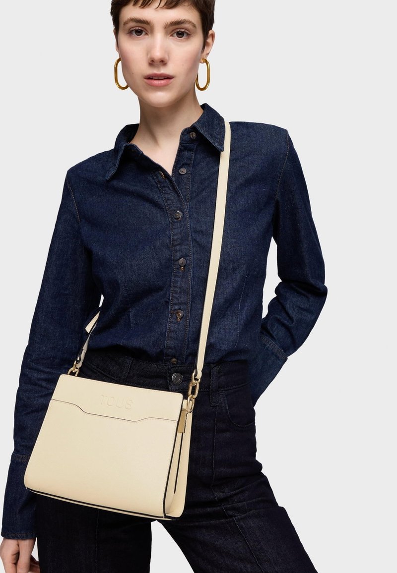Beżowa torba na ramię o teksturowanym wykończeniu, z klapą i złotymi akcentami. Noszona z ciemnoniebieską koszulą dżinsową.