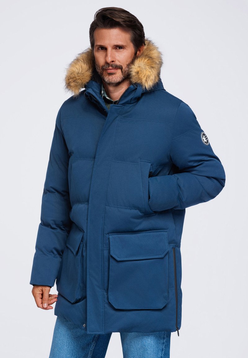 Homme portant un parka d'hiver bleu avec capuche bordée de fourrure et grandes poches avant, debout devant un fond clair uni.