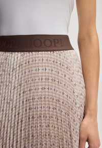 Mulher a usar uma saia plissada com um pequeno padrão castanho e uma cintura larga castanha com o texto "JOOP!", combinada com uma blusa branca.