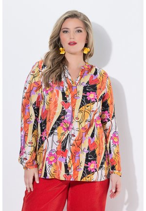 Frau mit langen blonden Haaren trägt eine bunte Bluse mit abstrakten weiblichen Gesichtern und roten Hosen, accessoriert mit gelben Blumenohrringen.
