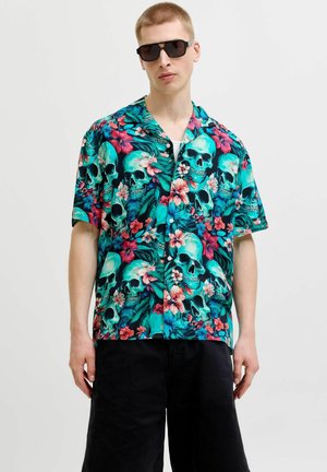 Jeune homme portant des lunettes de soleil noires et une chemise colorée à manches courtes avec un motif tête de mort et floral, associée à un short noir.